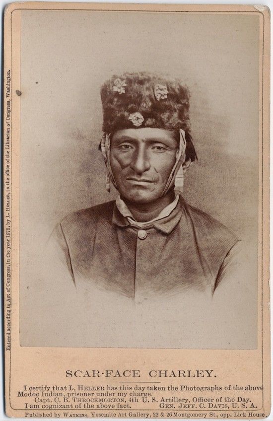A vintage photograph of a Native American man named Scar-Face Charley, wearing a fur hat and a dark coat, with prominent scars on his left cheek.