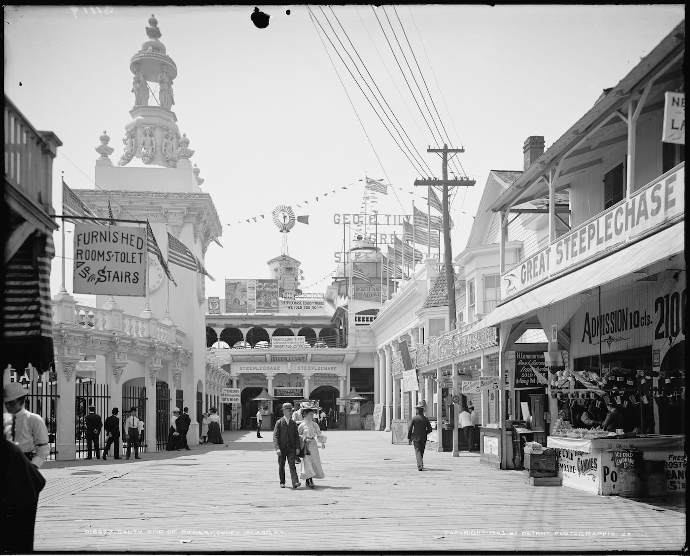 Black and white photograph of a crowded street on Coney Island, lined with buildings, signs, and people in period clothing.