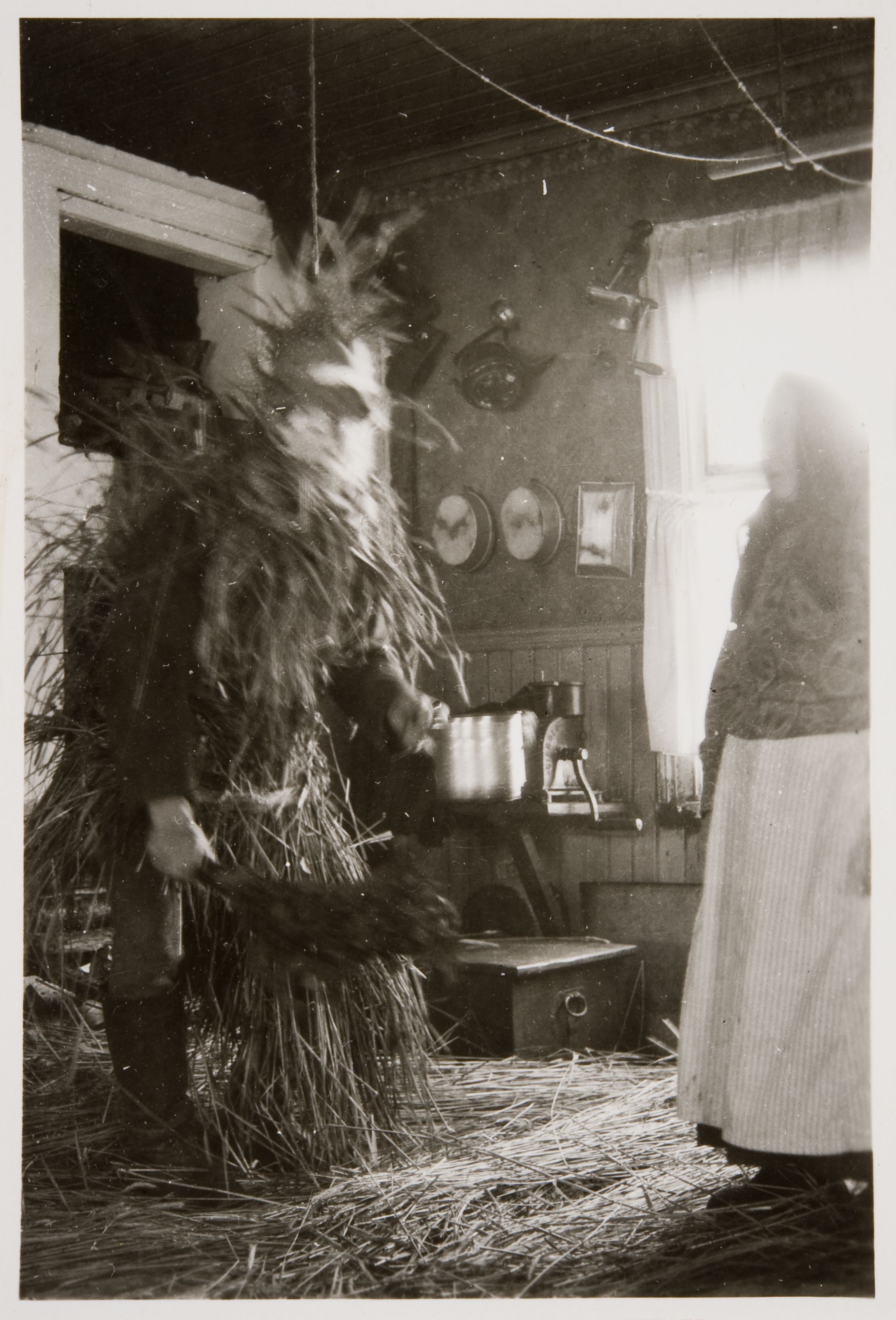 A vintage black and white photograph depicting a person in a straw costume in a rustic room.