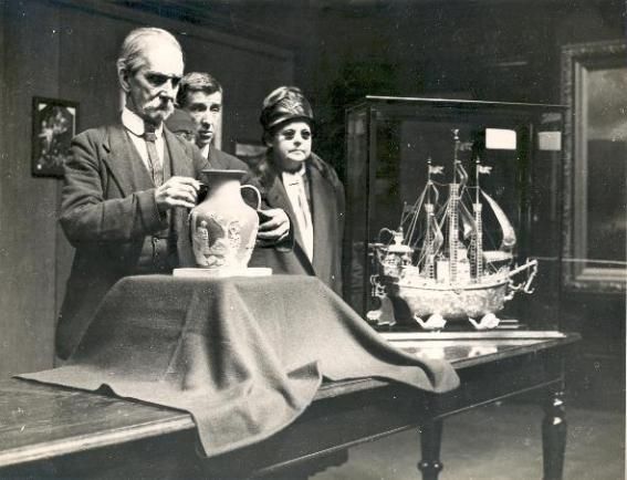 A black and white photograph of three people examining a vase and a ship model in a museum setting.