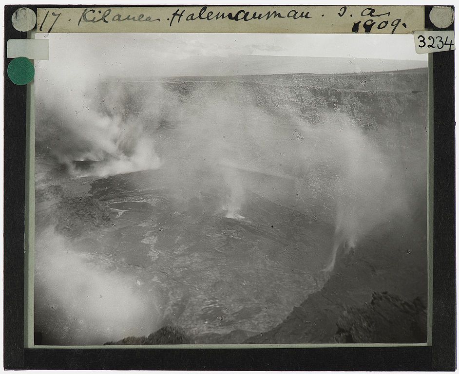 A vintage black and white photograph of a volcanic crater, likely Kilauea's Halemaumau, taken in 1909. The crater is filled with smoke and steam, and the surrounding landscape is rocky and barren.