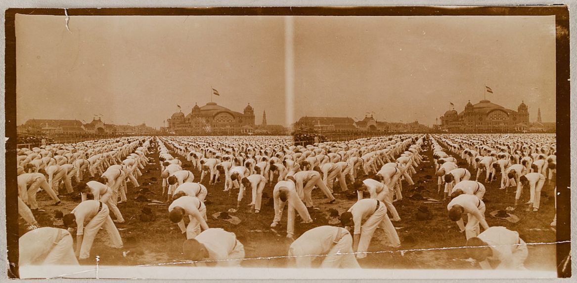 A sepia-toned photograph of a large group of people performing synchronized exercises in a vast field, with ornate buildings and flags in the background.