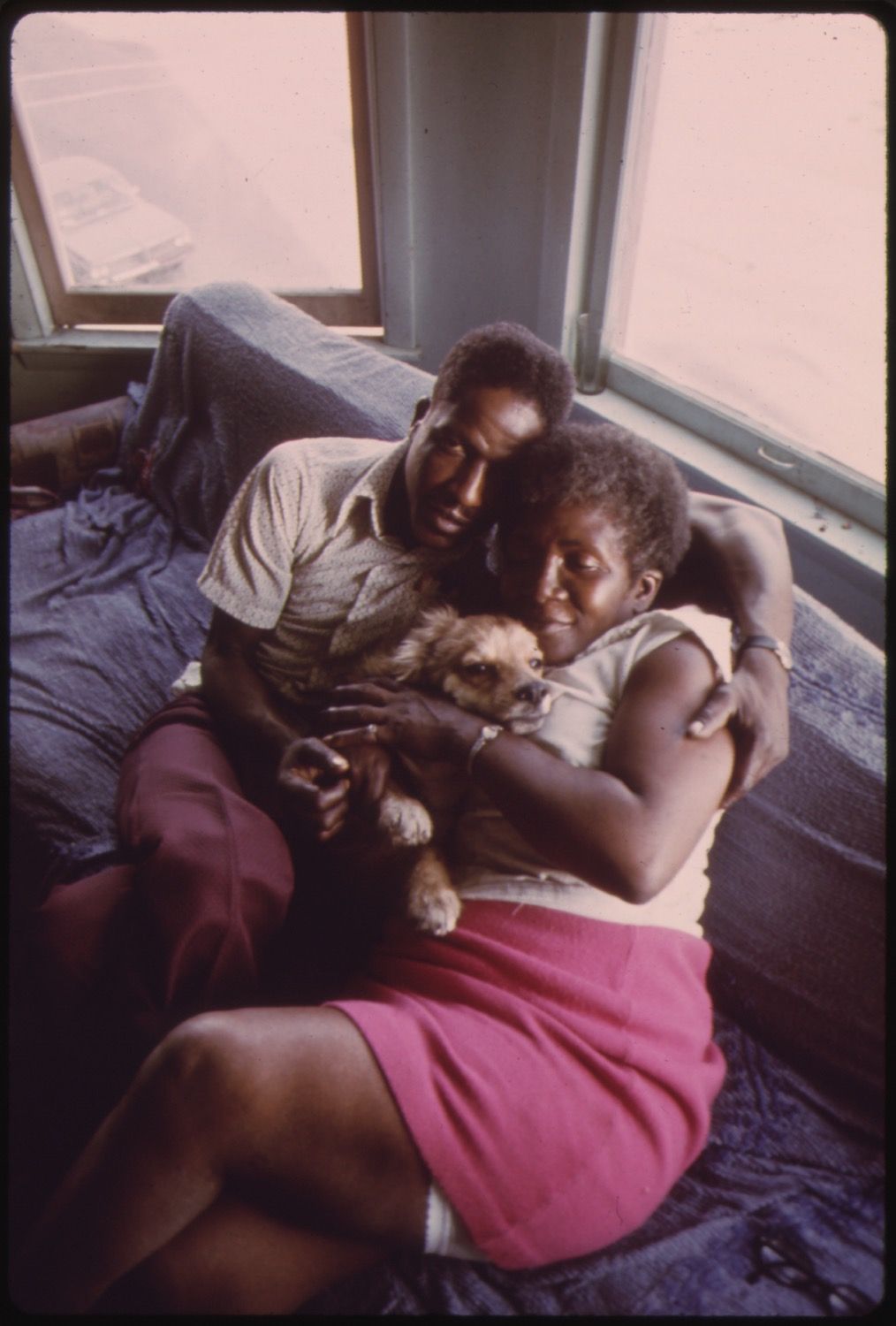 A Black couple sits on a blue couch, cradling a small, fluffy dog. The man is wearing a light-colored t-shirt and maroon pants. The woman is wearing a white tank top and a bright pink skirt. Behind them is a window with a view of a bright, hazy outdoor scene.
