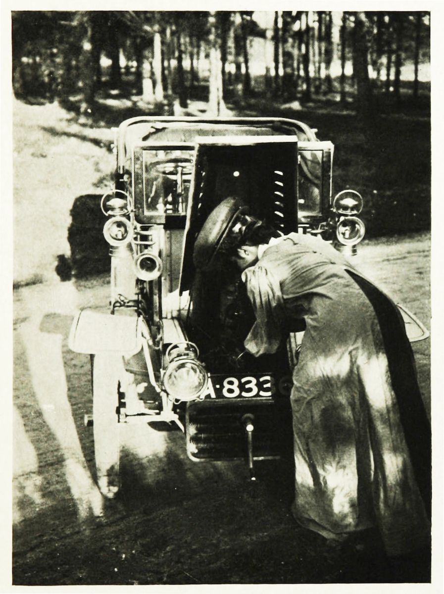 A vintage black and white photograph of a person working on the engine of an old car in a wooded area.