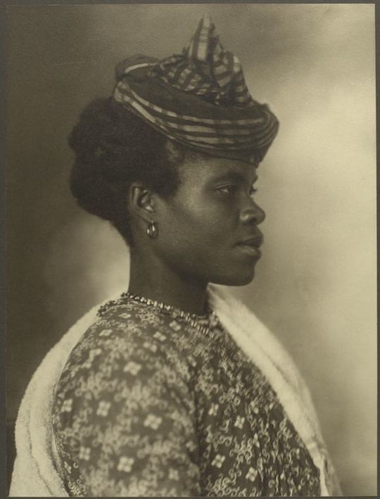 Sepia-toned portrait of a Black woman in profile, wearing a patterned hat and high-necked garment.