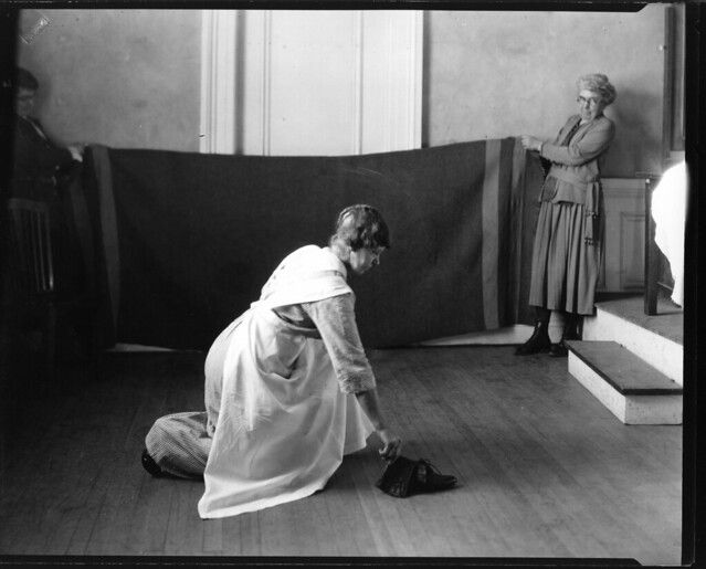A black and white photograph of two women in a room. One woman is kneeling and polishing shoes, while the other stands on stairs, observing her.