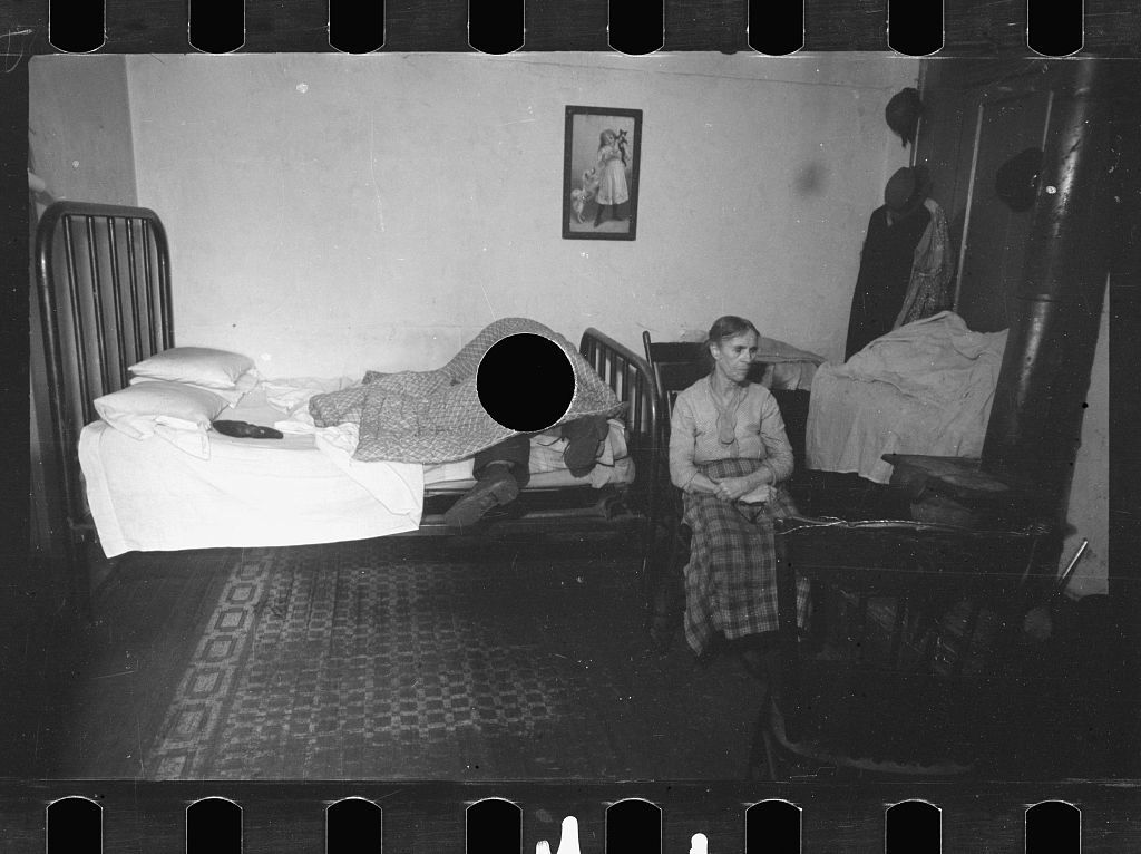 Black and white photograph of a room with two beds, a person sitting on one, and a child lying in the other.