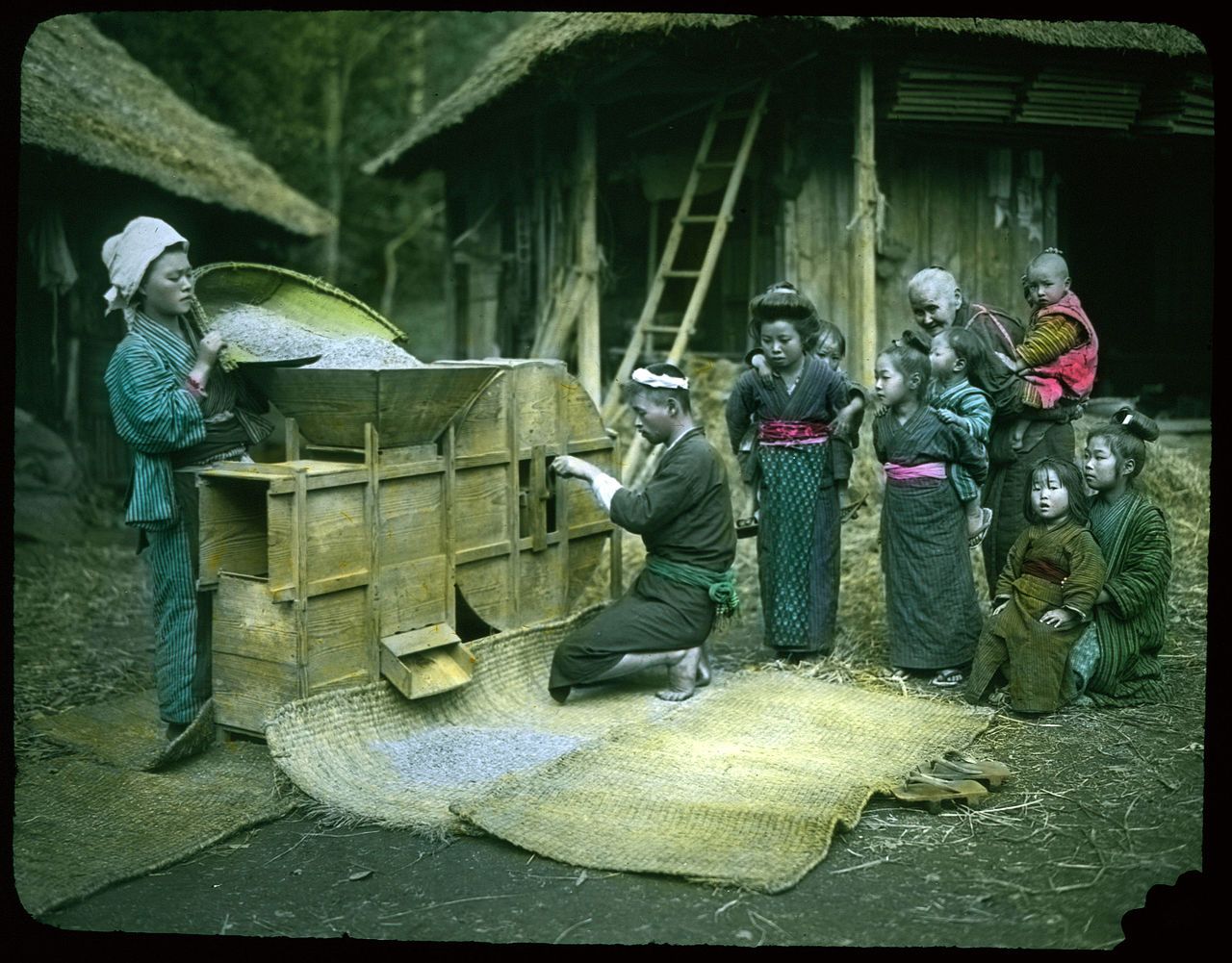 A group of people are gathered around a threshing machine in a rural Japanese setting.