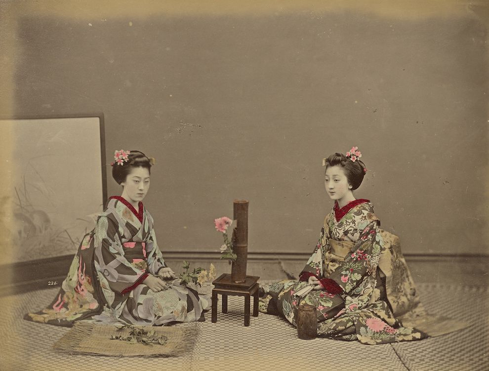 Vintage photograph of two Japanese women in kimonos seated on a tatami mat floor in a traditional room.