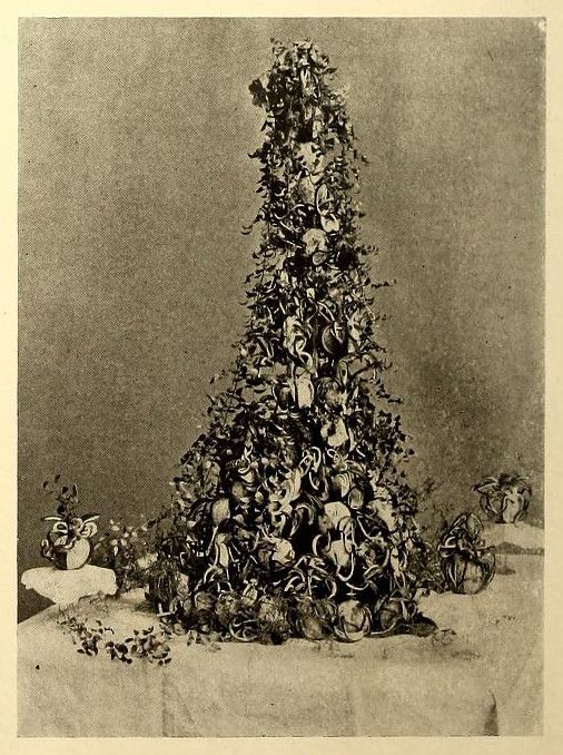 Vintage black and white photograph of a Christmas tree densely decorated with ornaments, standing on a table with figures on either side.
