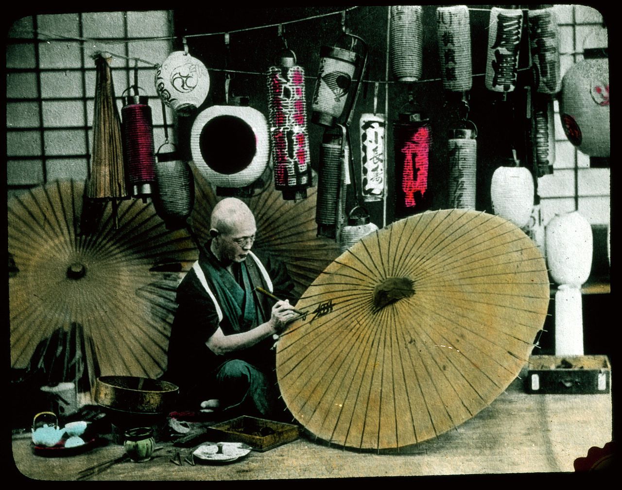 A vintage-style photograph of a craftsman working on a traditional Japanese umbrella in a workshop filled with colorful lanterns.