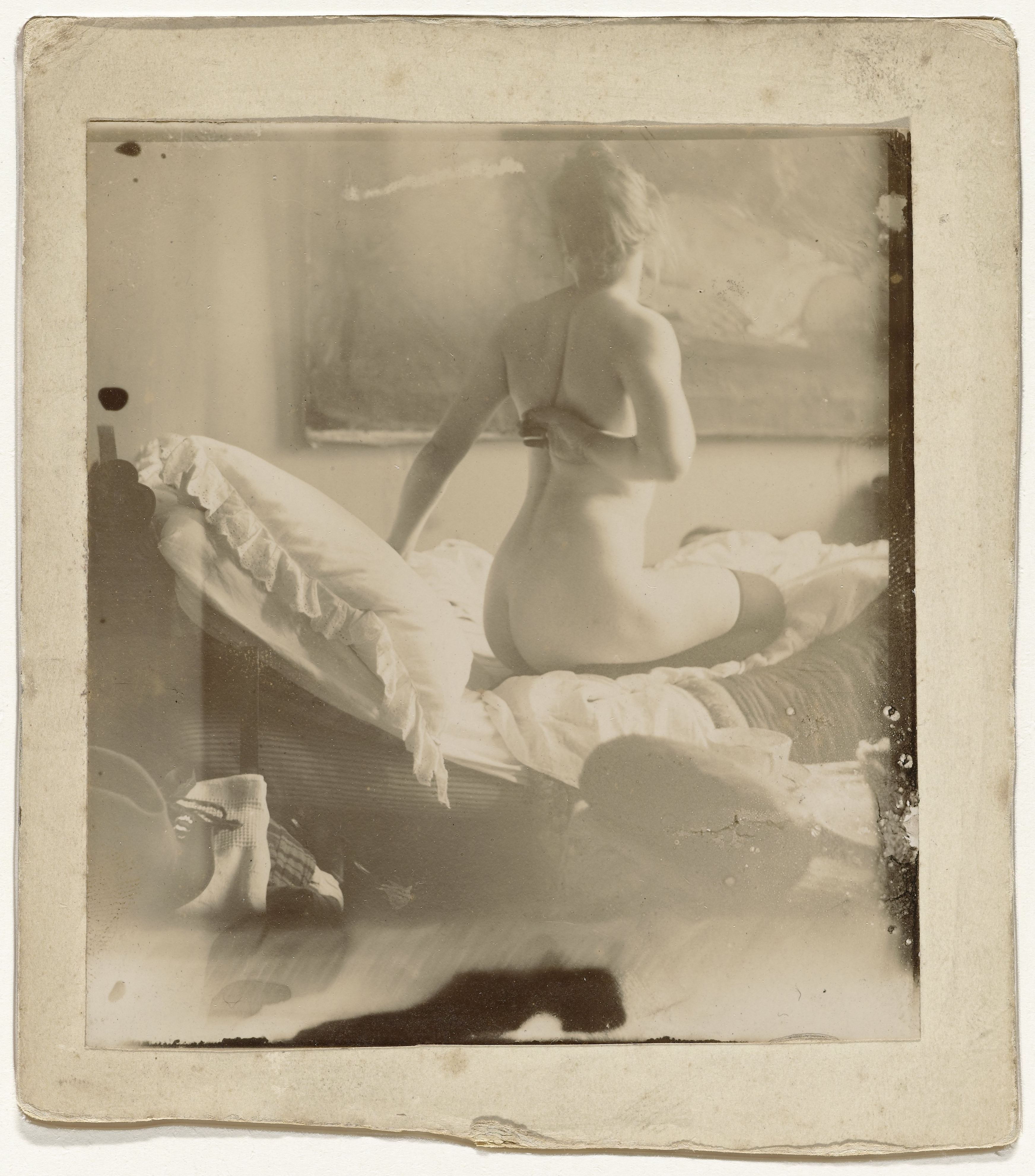 Vintage sepia-toned photograph of a nude woman seated on a bed, draped in white fabric and pillows.