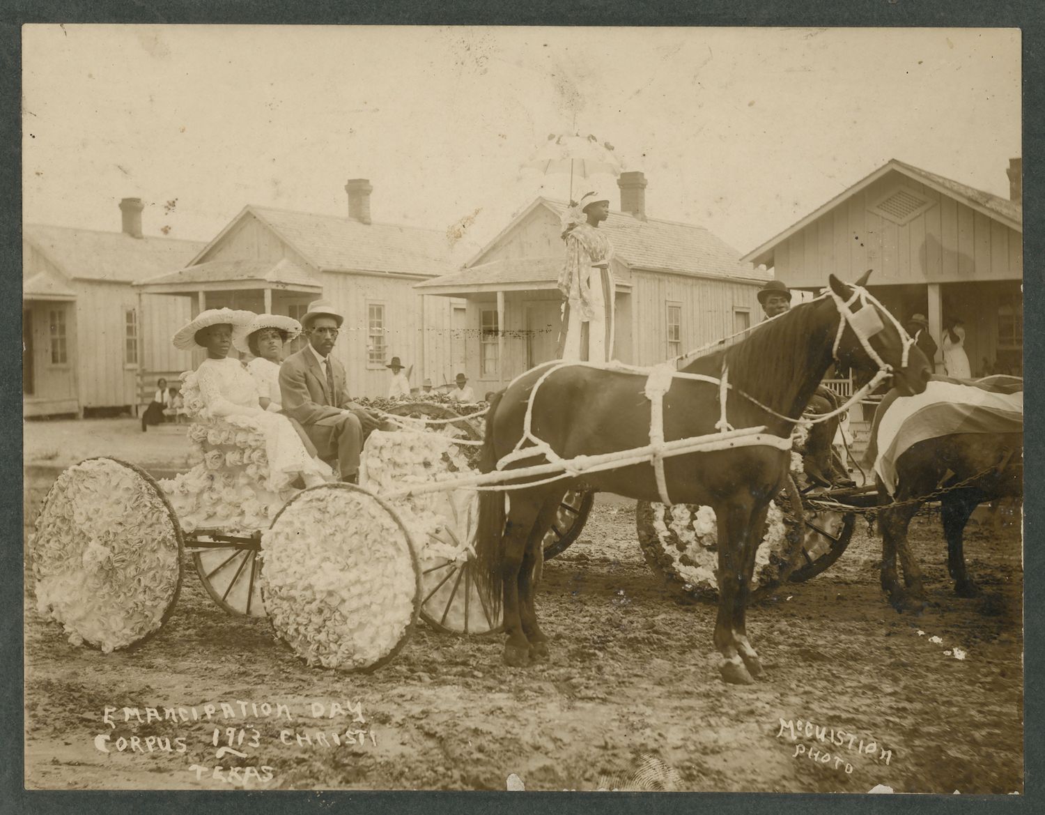 A sepia-toned photograph from 1913, taken in Corpus Christi, Texas, commemorating Emancipation Day. The scene depicts a horse-drawn carriage or wagon decorated with what appears to be cotton. In the carriage, there are three African American individuals. The person in the center is a man wearing a suit and hat. To his left is a woman wearing a dress and a large hat, and to his right is another woman, also in a dress and hat. The horse is dark-colored and harnessed to the wagon. A man is seen standing near the horse, possibly the driver or caretaker. The background shows a street lined with simple wooden buildings, likely homes or businesses. There are a few trees and a fence visible as well.