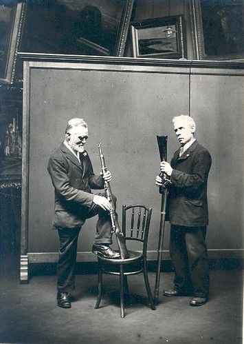 Two men in suits stand with long musical instruments in front of a paneled wall.