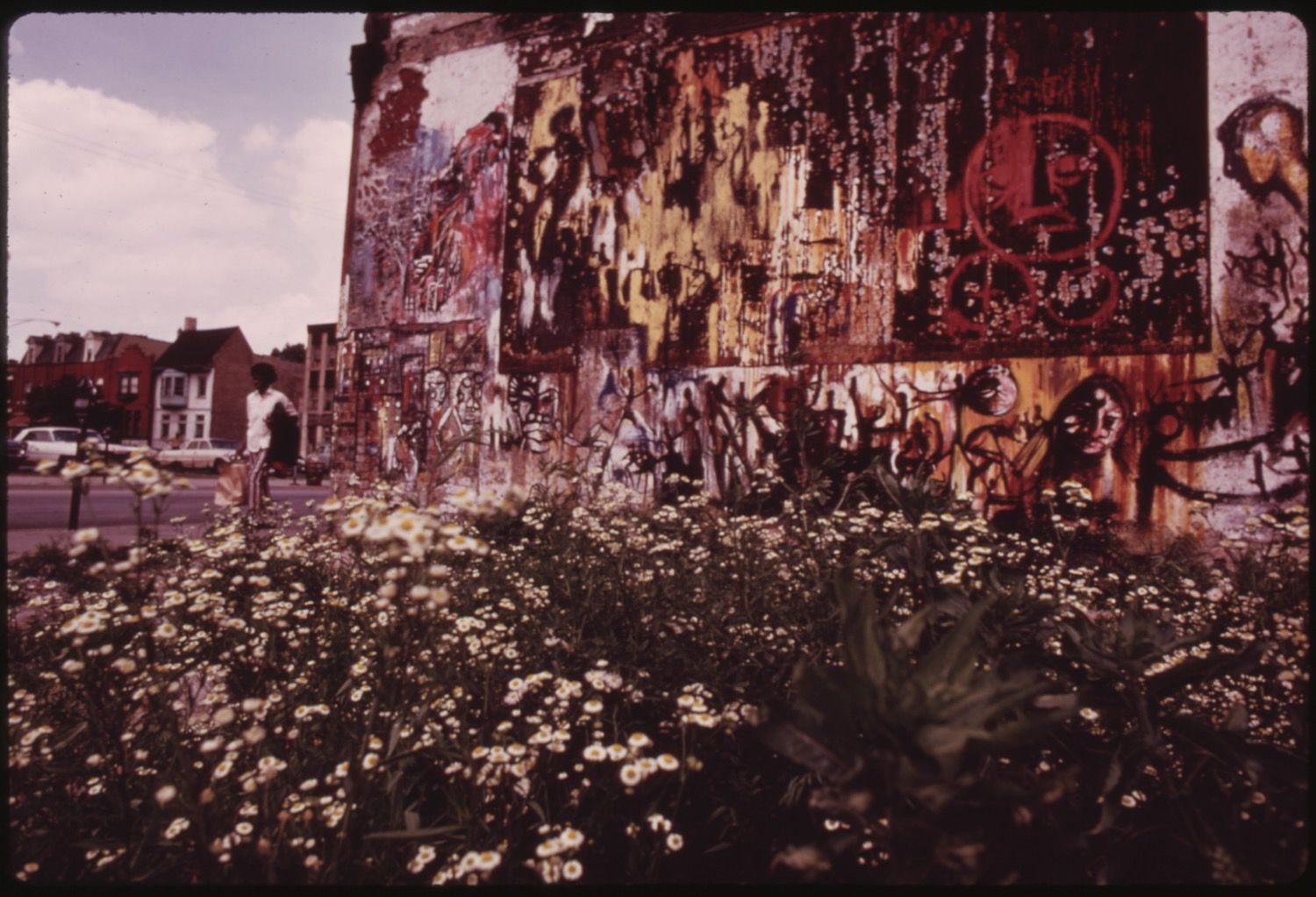 A weathered wall covered in layers of faded posters and graffiti, with a dense patch of wildflowers growing at its base. Two people are walking along the street nearby.