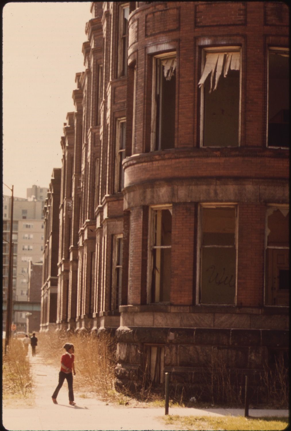 A row of tall, brick buildings with a sense of decay and abandonment. A person is walking away from the viewer on the sidewalk.