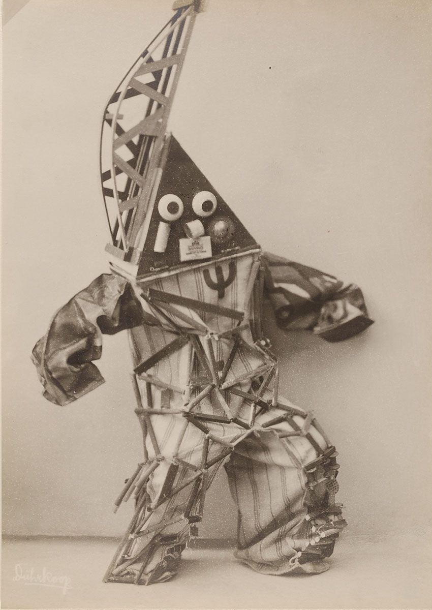 A black and white photograph of an abstract sculpture resembling a human figure constructed from metal wires, fabric scraps, and paper collage elements.