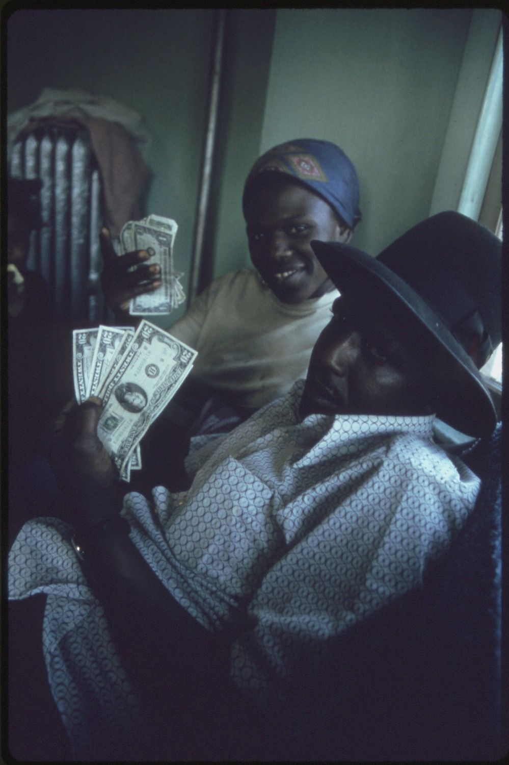 A woman and a child in a living room. The woman is holding a fan of US currency, displaying it with a smile. The child is lying on a couch, partially obscured by a patterned blanket.