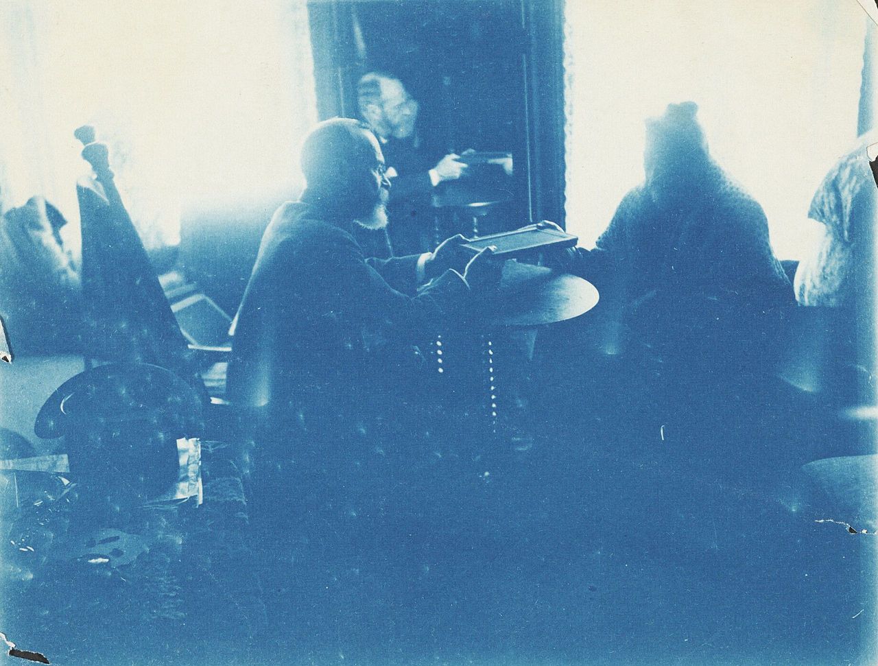 A vintage cyanotype photograph of a man reading at a table in a room with other people.