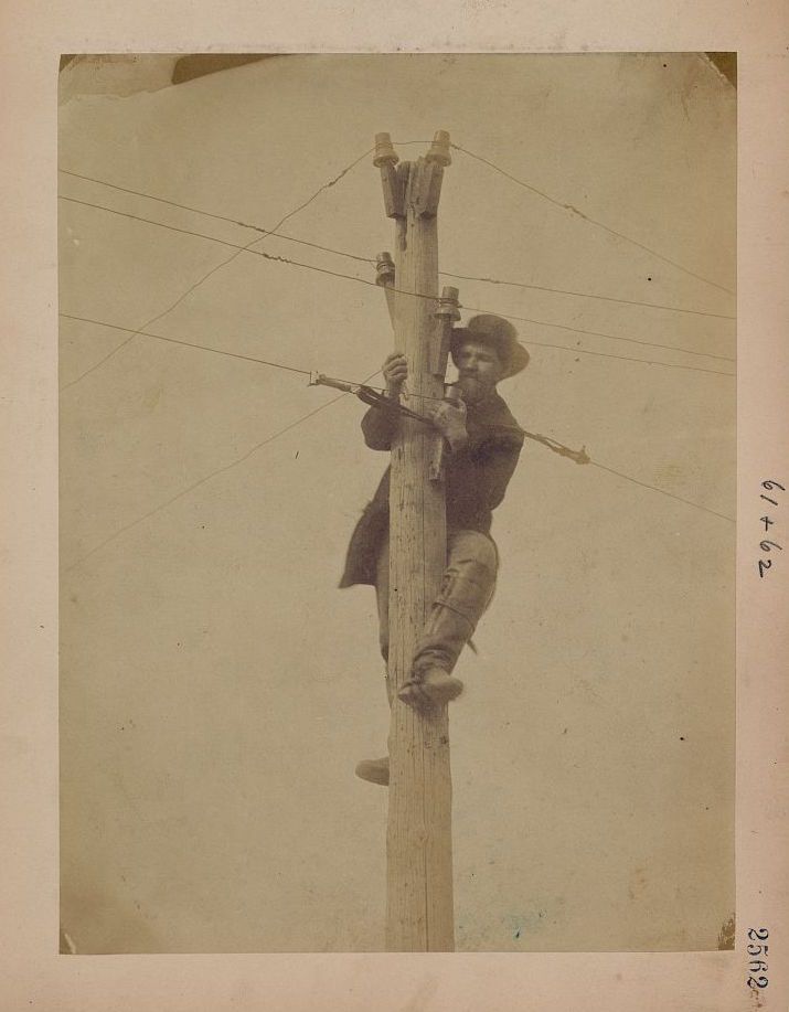 Vintage photograph of a man climbing a wooden utility pole, working with wires and tools.