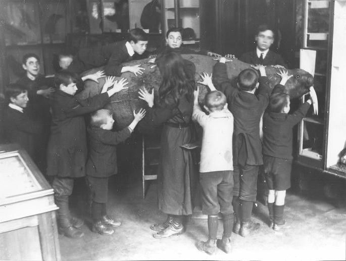 A black and white vintage photograph of a woman surrounded by children in a classroom or library setting. The children are reaching out and seemingly 'holding' or supporting a large, circular object that appears to be draped around her.