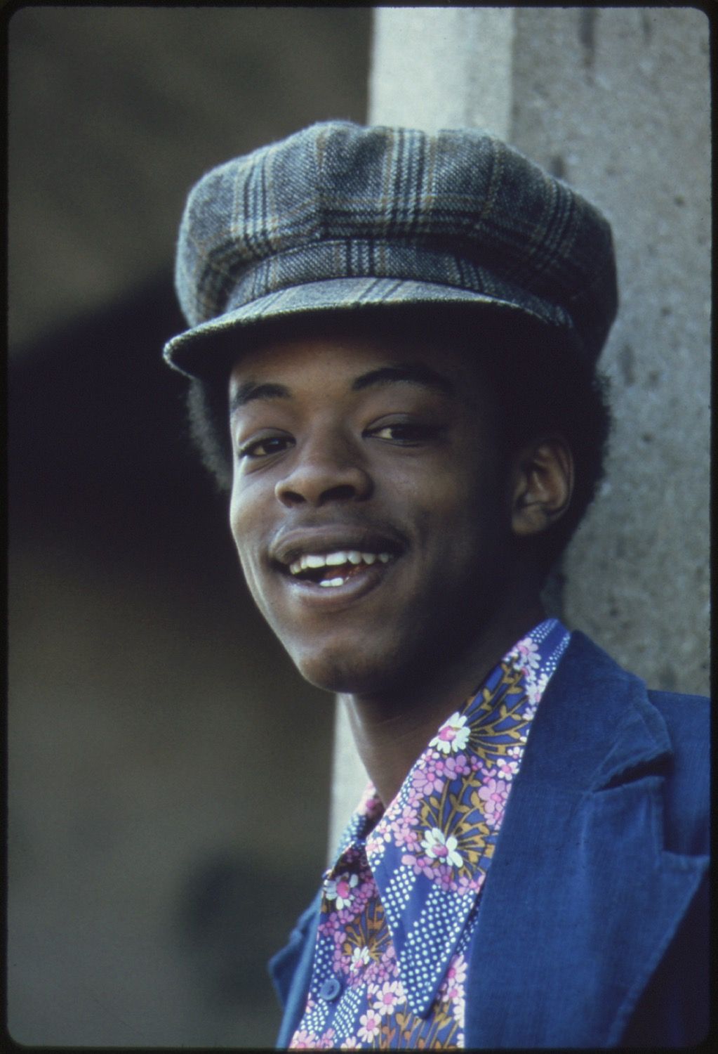A portrait of a young Black man wearing a tweed newsboy cap, floral shirt, and blue jacket, smiling warmly at the camera against a concrete wall.