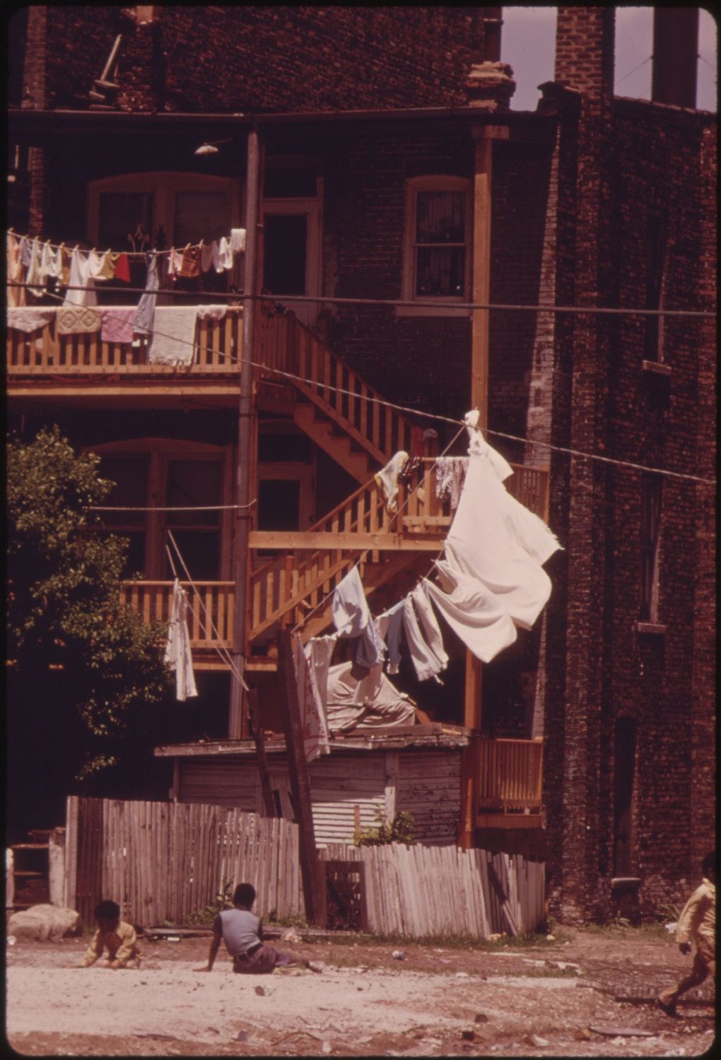 A weathered, multi-story wooden building with laundry hanging from lines strung between it and a nearby structure. Two children are visible in the foreground.