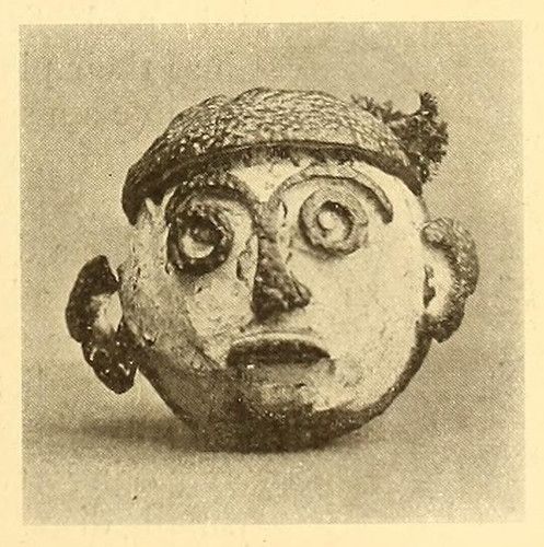 Close-up of a weathered mask with a distorted face and textured headdress.