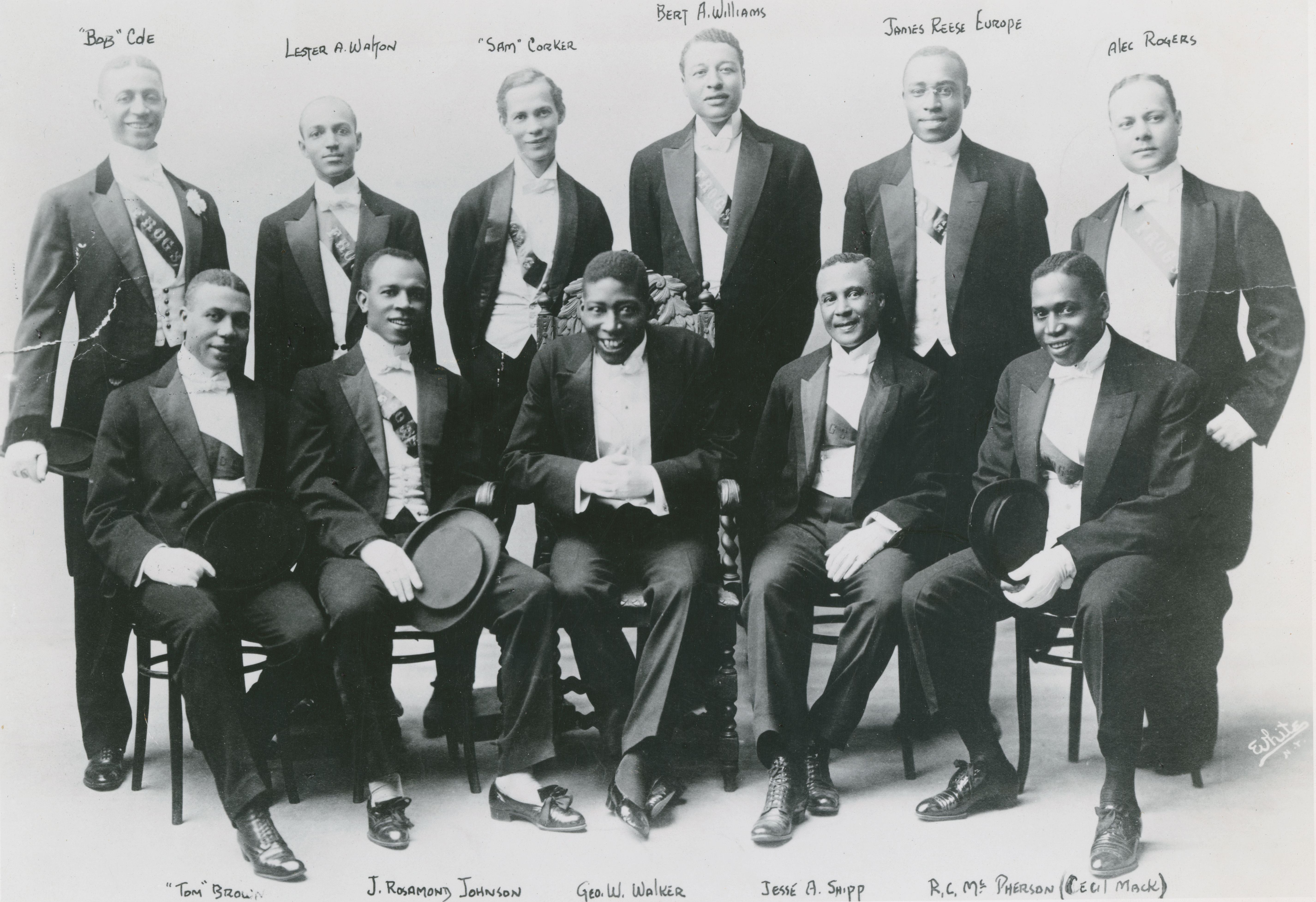 A black and white photograph of thirteen formally dressed men, identified by names below them, arranged in a tiered formation.