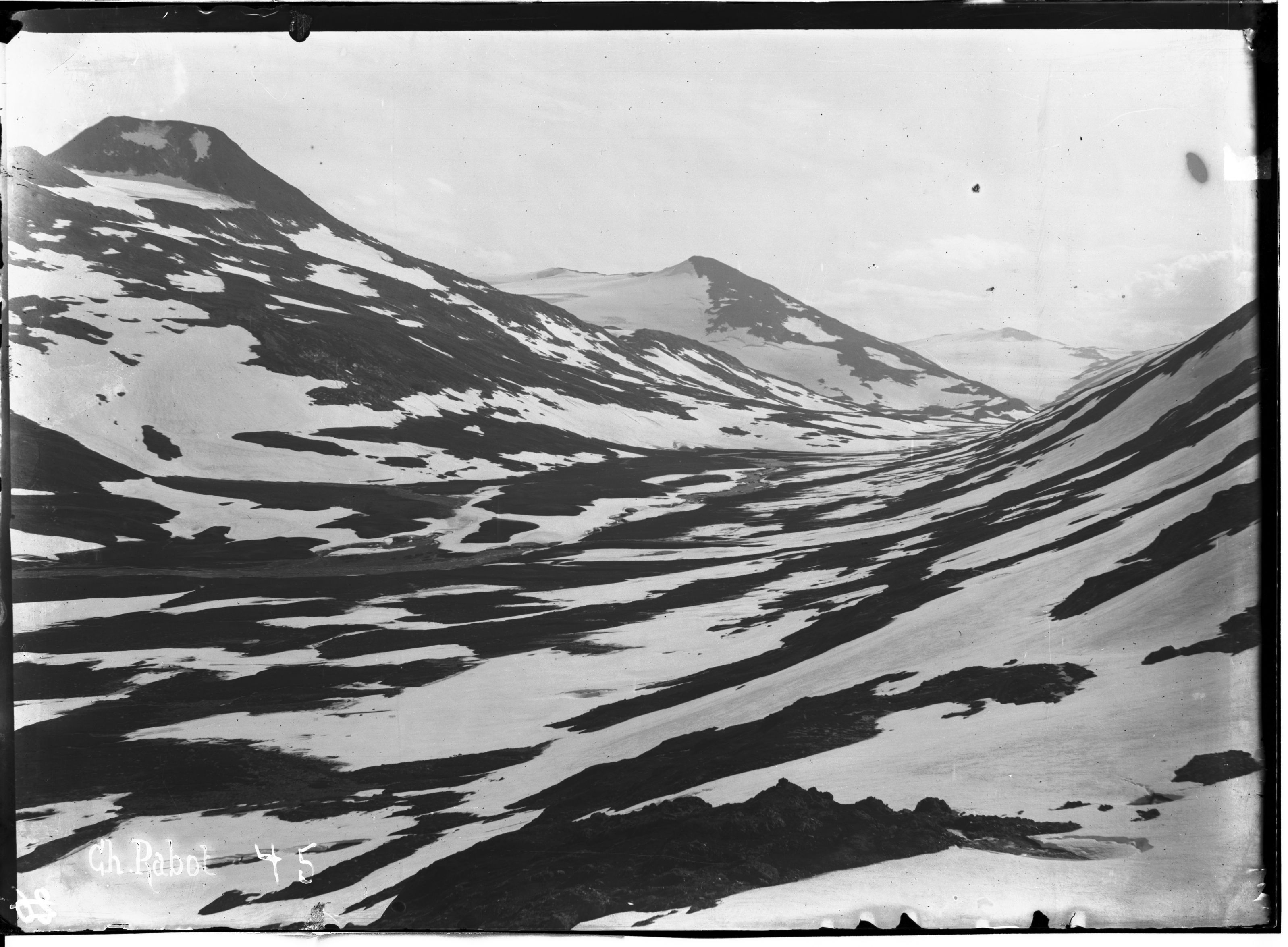 A black and white photograph of a mountainous valley with snow-covered peaks and dramatic shadows.