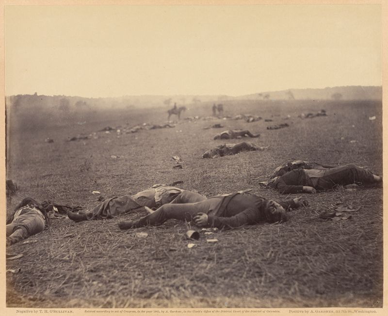 A sepia-toned photograph of a battlefield covered in the bodies of fallen soldiers. The scene is a vast, grassy field with trees and a hazy sky in the distance. A lone horse and rider can be seen in the far background.