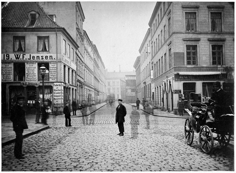 A black and white photograph of a cobblestone street lined with tall buildings in a European city. People are walking and standing on the street, and a horse-drawn carriage is visible on the right. Shopfronts with signs and advertisements line the street.