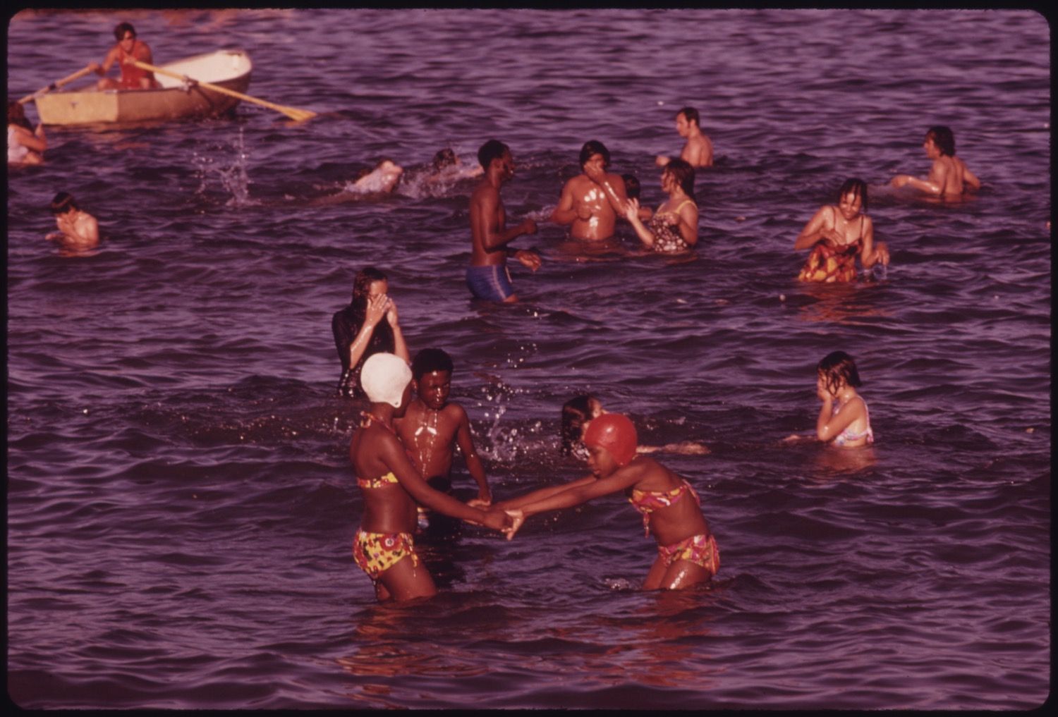 A group of people swimming in a body of water, with two young men playfully interacting in the foreground and a small boat in the background.