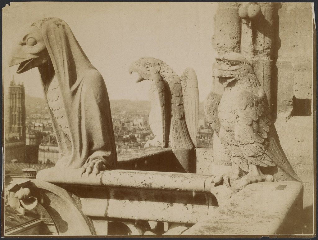 A sepia-toned photograph of stone gargoyles on a building, with a cityscape in the background.