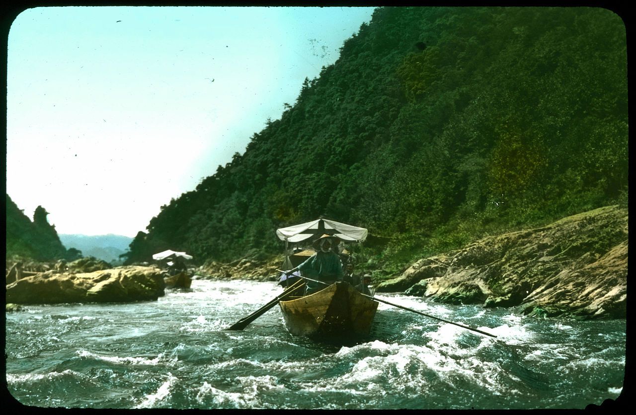 Vintage color photograph of two small boats navigating a fast-flowing river with a forested mountain in the background.