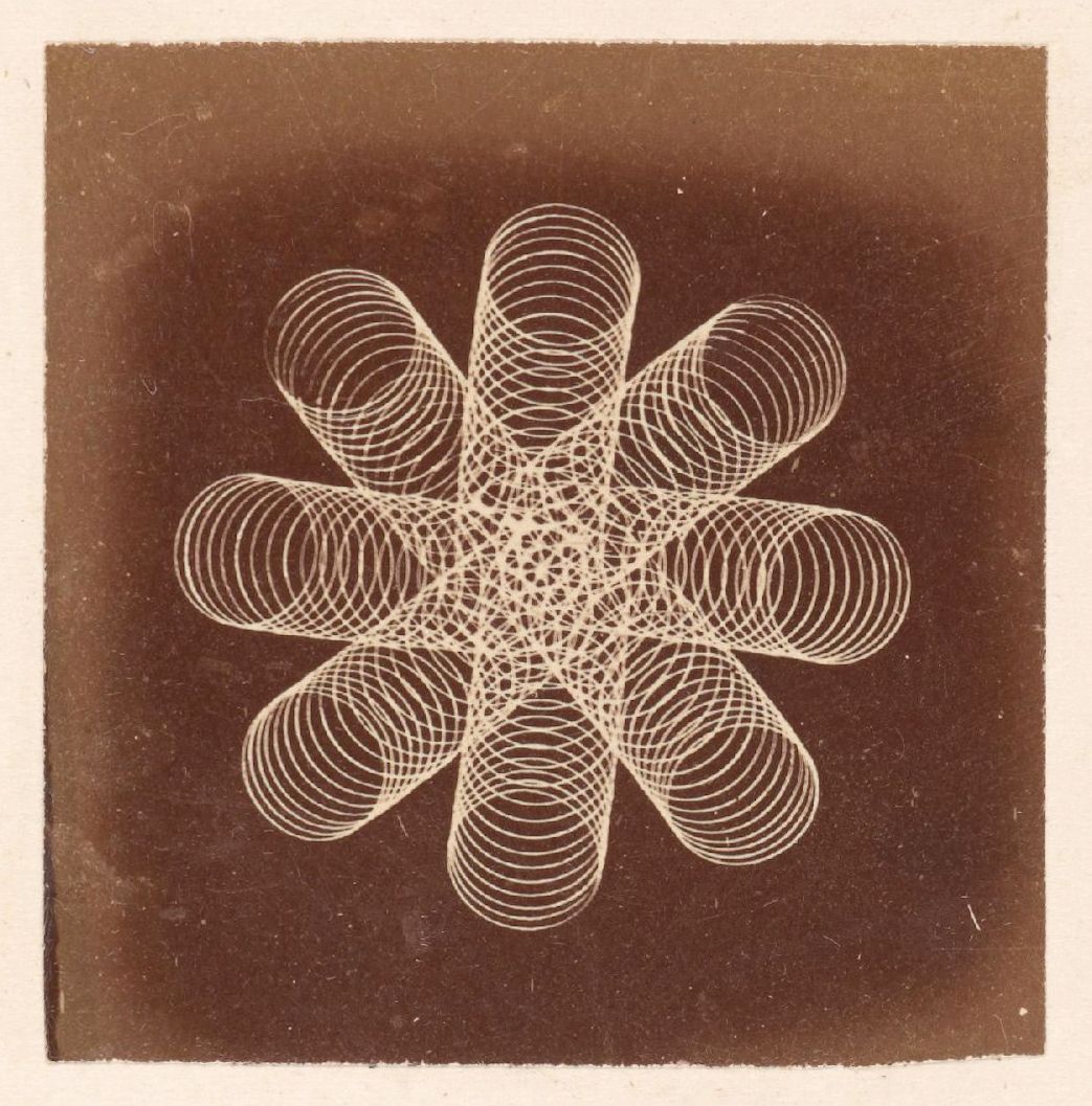 A vintage photograph of a complex geometric pattern resembling a flower or starburst, composed of spiraling lines against a dark background.