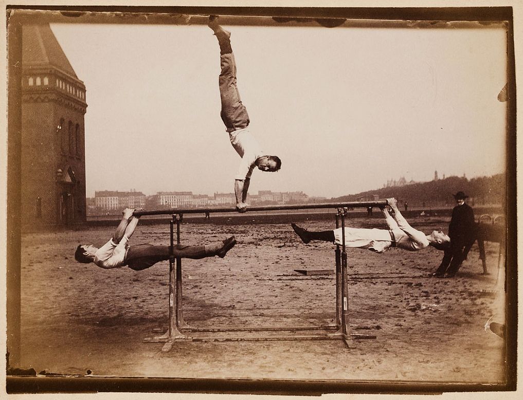 A sepia-toned photograph of three gymnasts performing on parallel bars in an outdoor setting. One gymnast is in mid-air, while the other two are suspended from the bars. A man stands nearby, watching. A cityscape and a building with a turret are visible in the background.