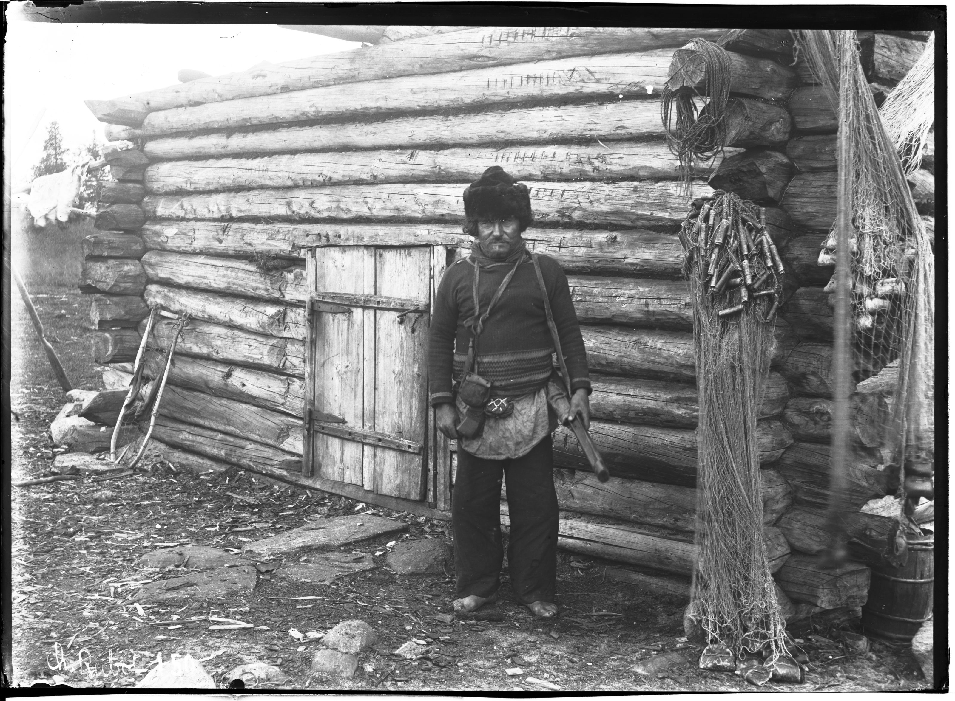 A black and white photograph of a man standing in front of a log cabin.