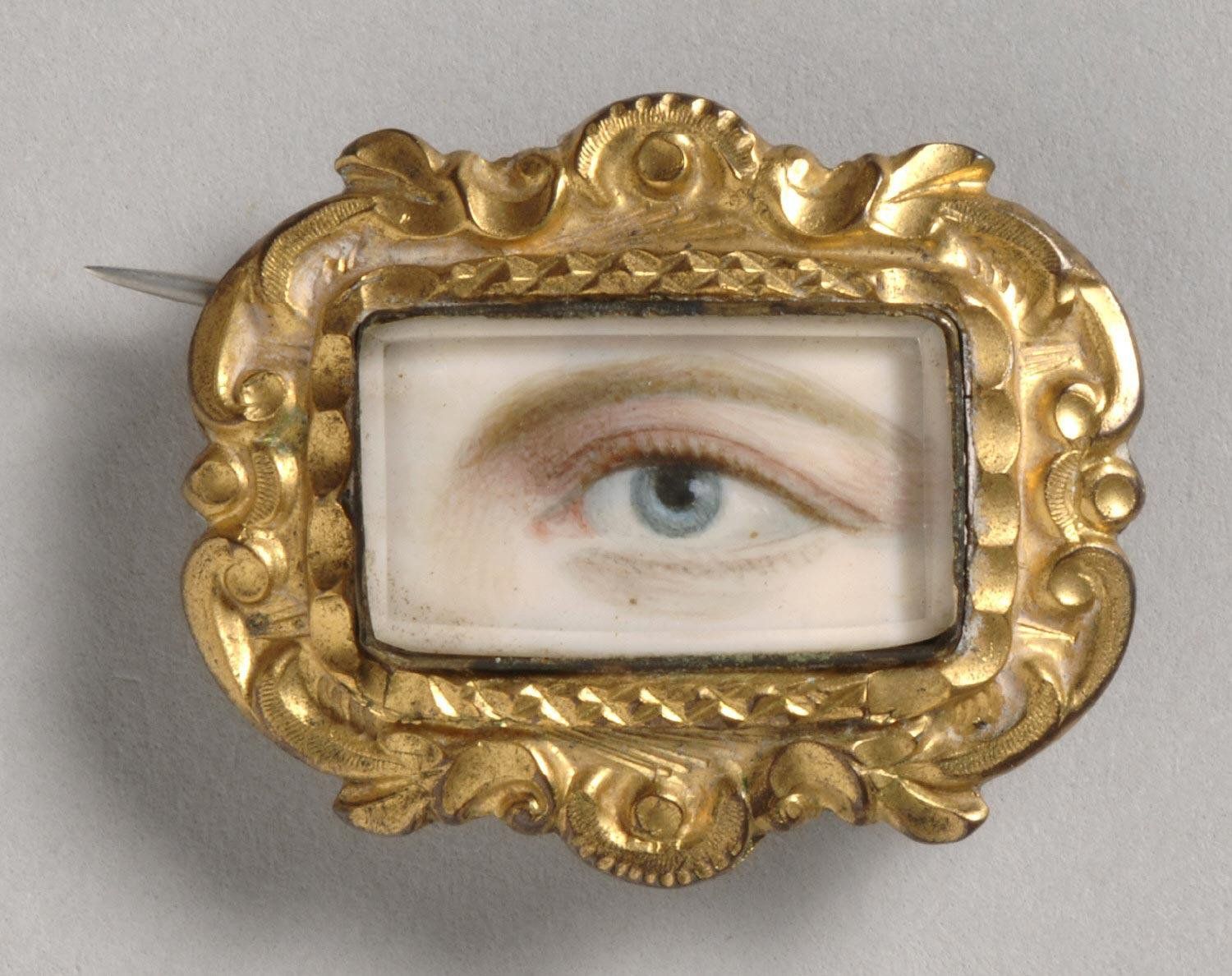 Close-up of a vintage brooch featuring a realistic painting of a blue human eye within an ornate, gilded frame.