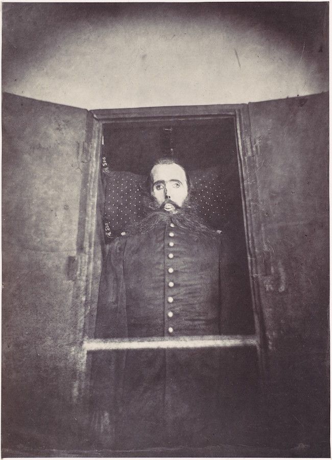 A black and white photograph of a man framed within a doorway, with a weathered wall surrounding him.