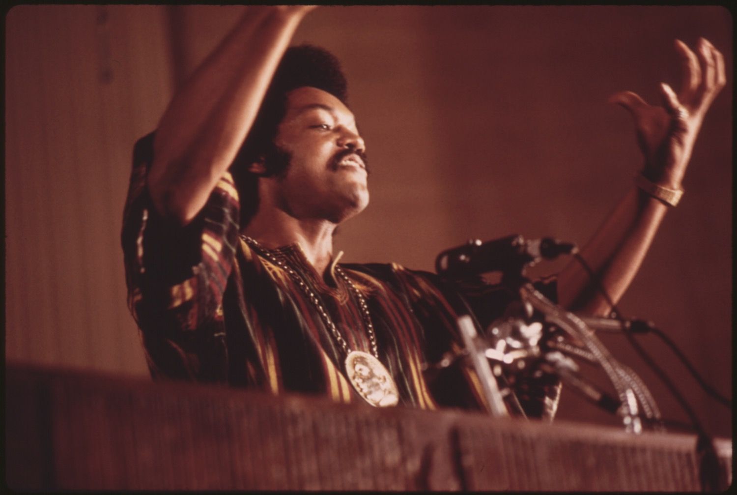 Close-up of a man on stage with a microphone, wearing a patterned shirt and a large pendant, with his arm raised in a gesture.