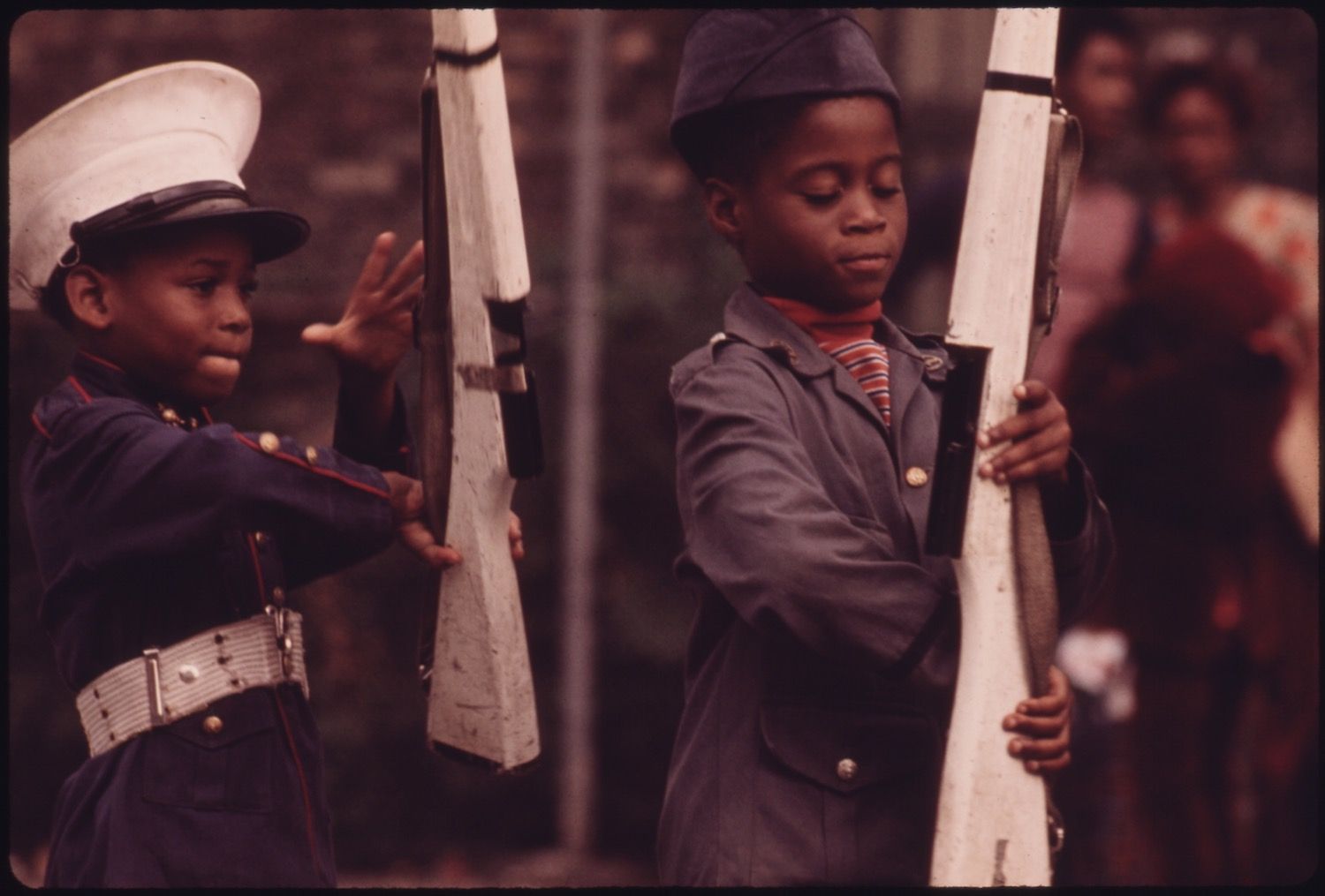Two young boys in military uniforms holding white rifles, with a blurred crowd in the background.
