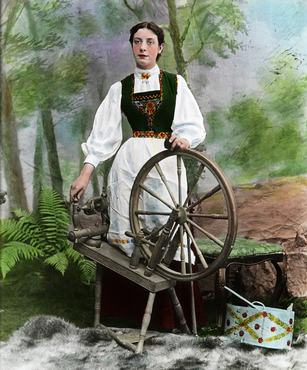 A hand-colored photograph of a young woman standing next to a spinning wheel in a forest setting.