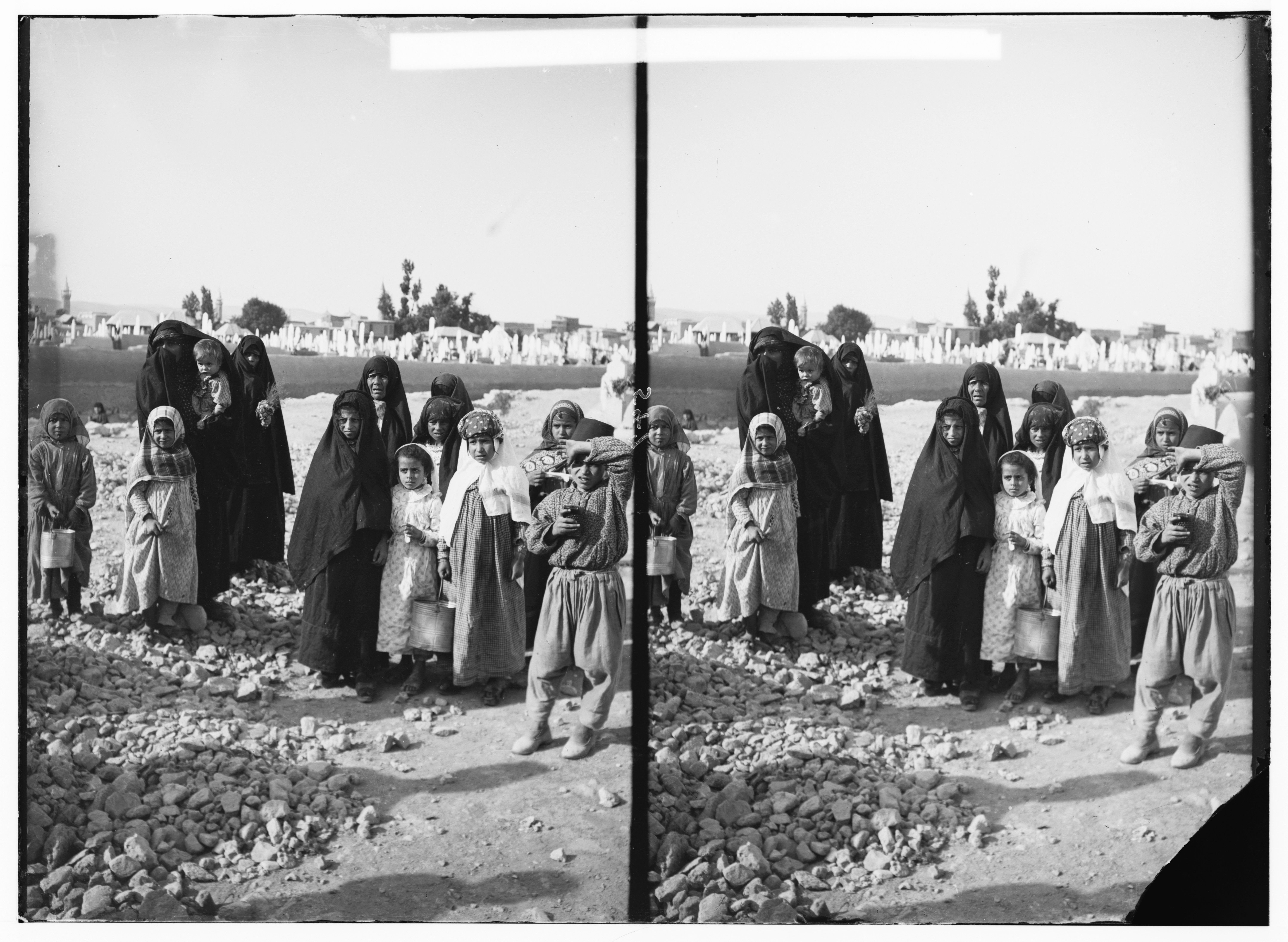 A black and white stereoscopic photograph of a group of people, including women and children, standing in a rubble-strewn area.