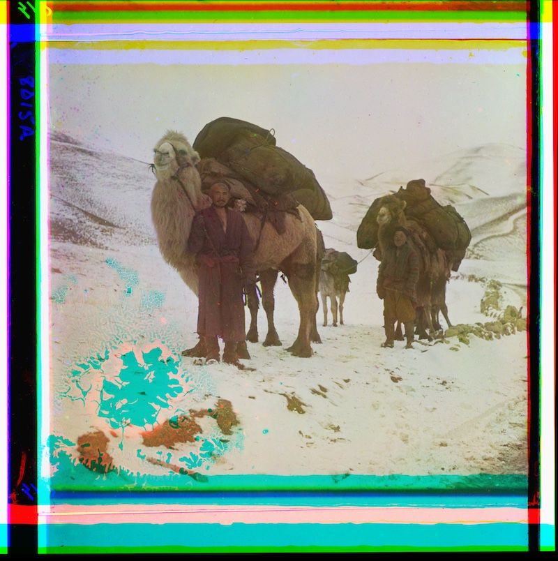 A line of camels carrying large bundles across a snowy landscape, with mountains in the background and a colorful border around the image.