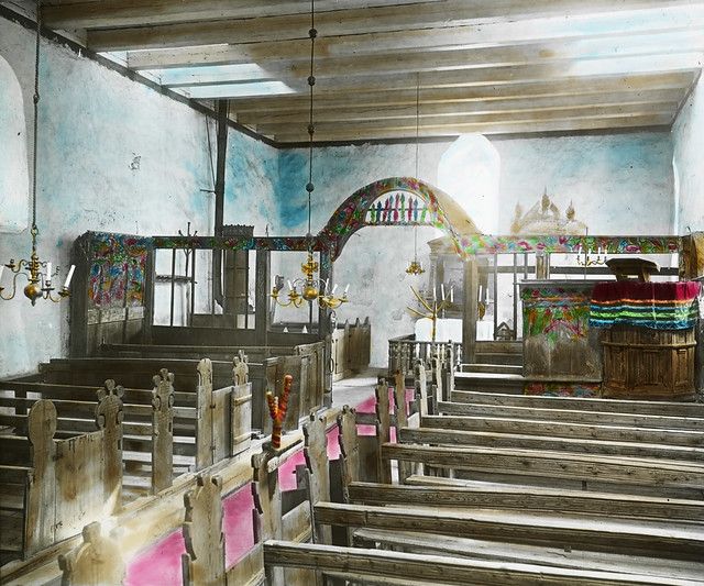 Interior of a historic synagogue with wooden benches, colorful decorations, and a chandelier.