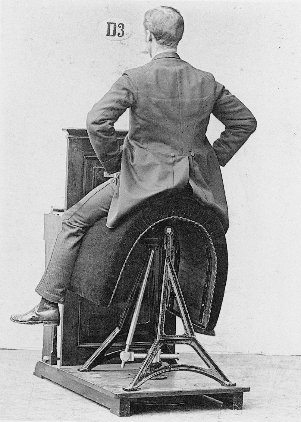 Black and white photograph of a man seated on a complex machine, possibly a therapeutic device, with a number 'D3' in the top left corner.