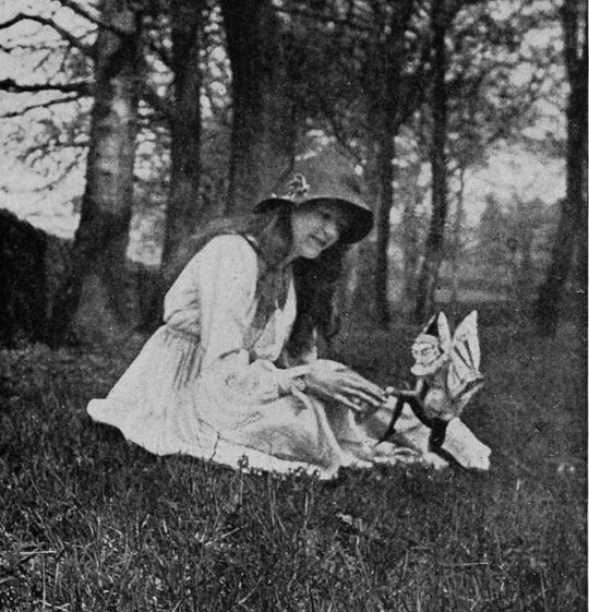 A vintage black and white photograph of a young girl in a dress and hat interacting with a fairy figurine in a forest.