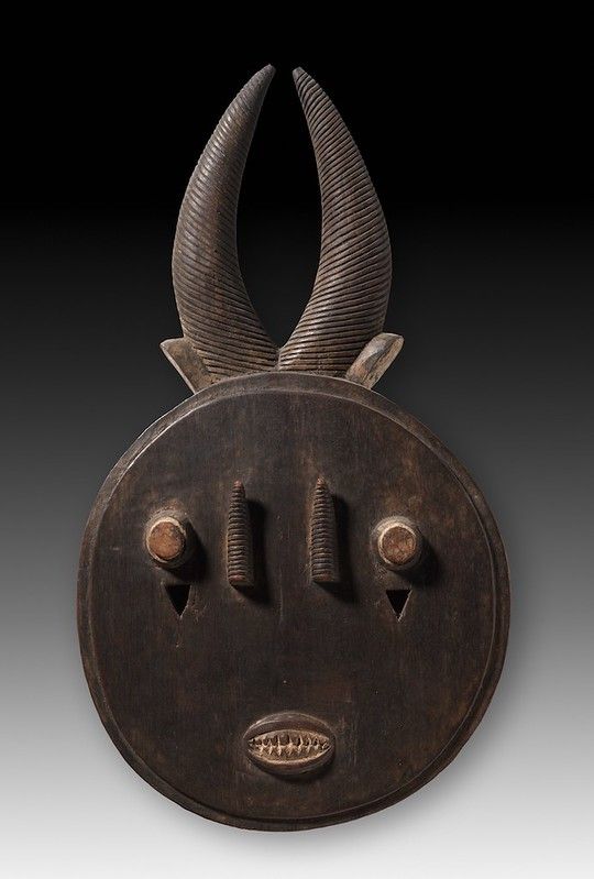A dark, wooden African mask with curved horns, stylized eyes, nostrils, and a carved mouth.