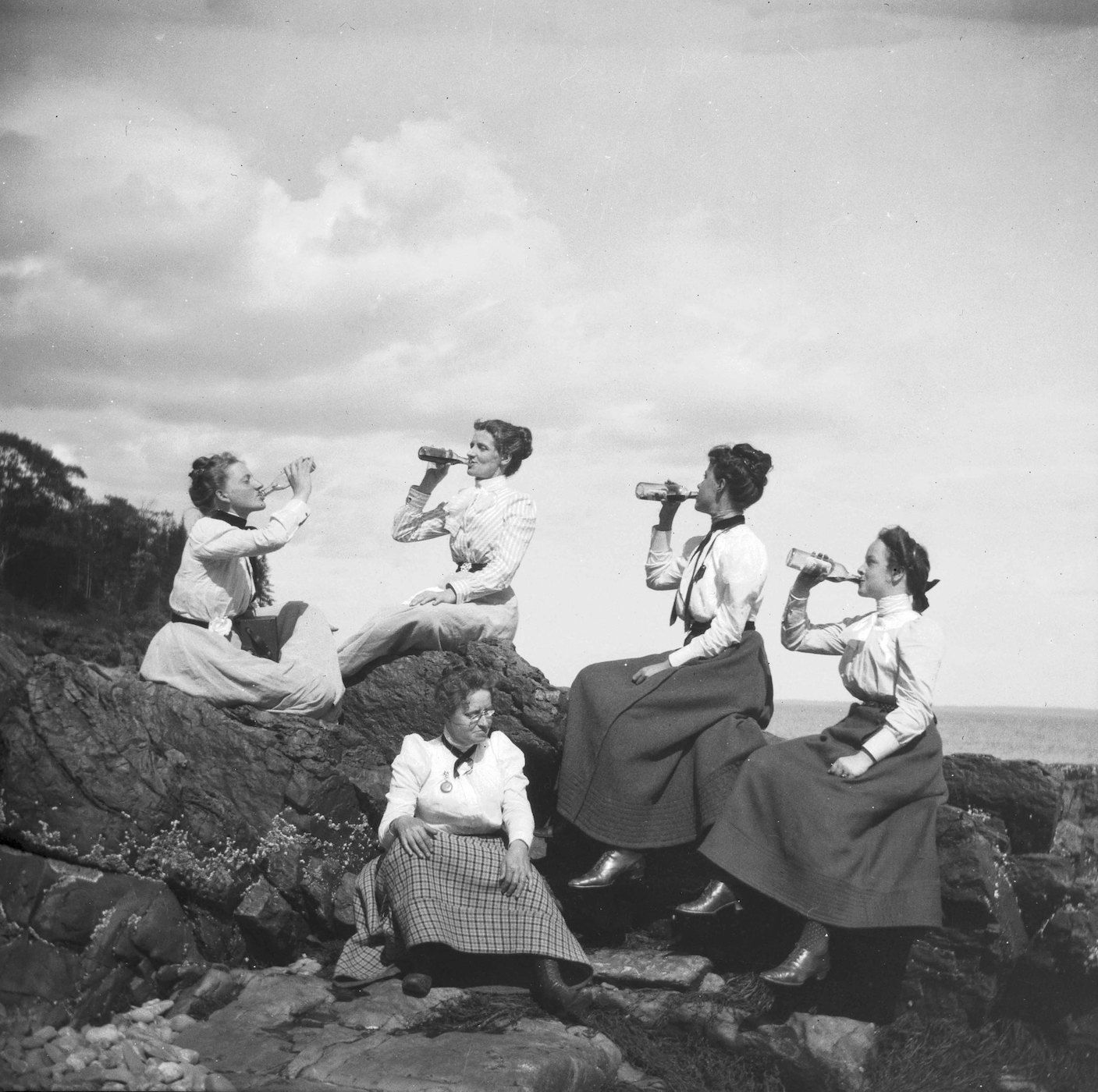 A black and white photograph of five women drinking from bottles while sitting on a rocky cliffside.